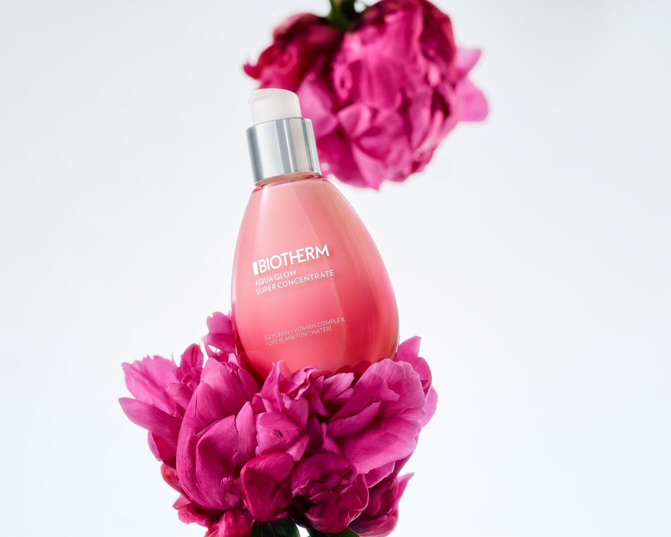 Rožinis „Biotherm Aqua Glow Super Concentrate“ buteliukas su sidabriniu dozatoriumi, apsuptas ryškiai rožinių žiedų, pabrėžiančių produkto švytėjimą suteikiančias savybes.