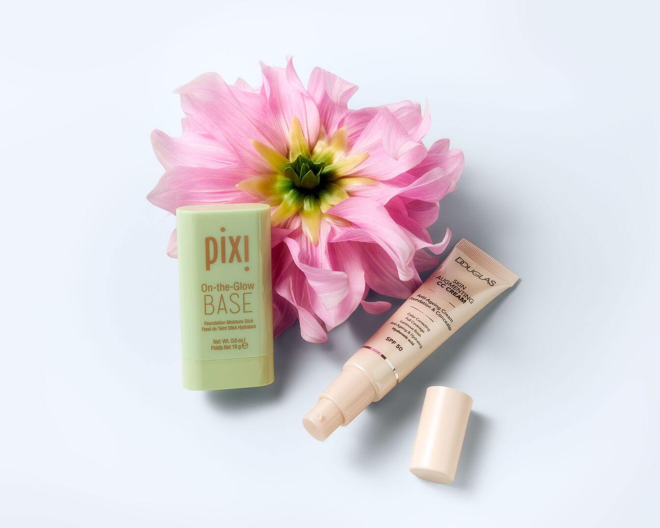 Šviesiai žalios spalvos „Pixi On-the-Glow BASE“ pagrindo lazdelė šalia rausvo žiedo ir „Douglas Skin Augmenting CC Cream“ kremo su SPF 50.