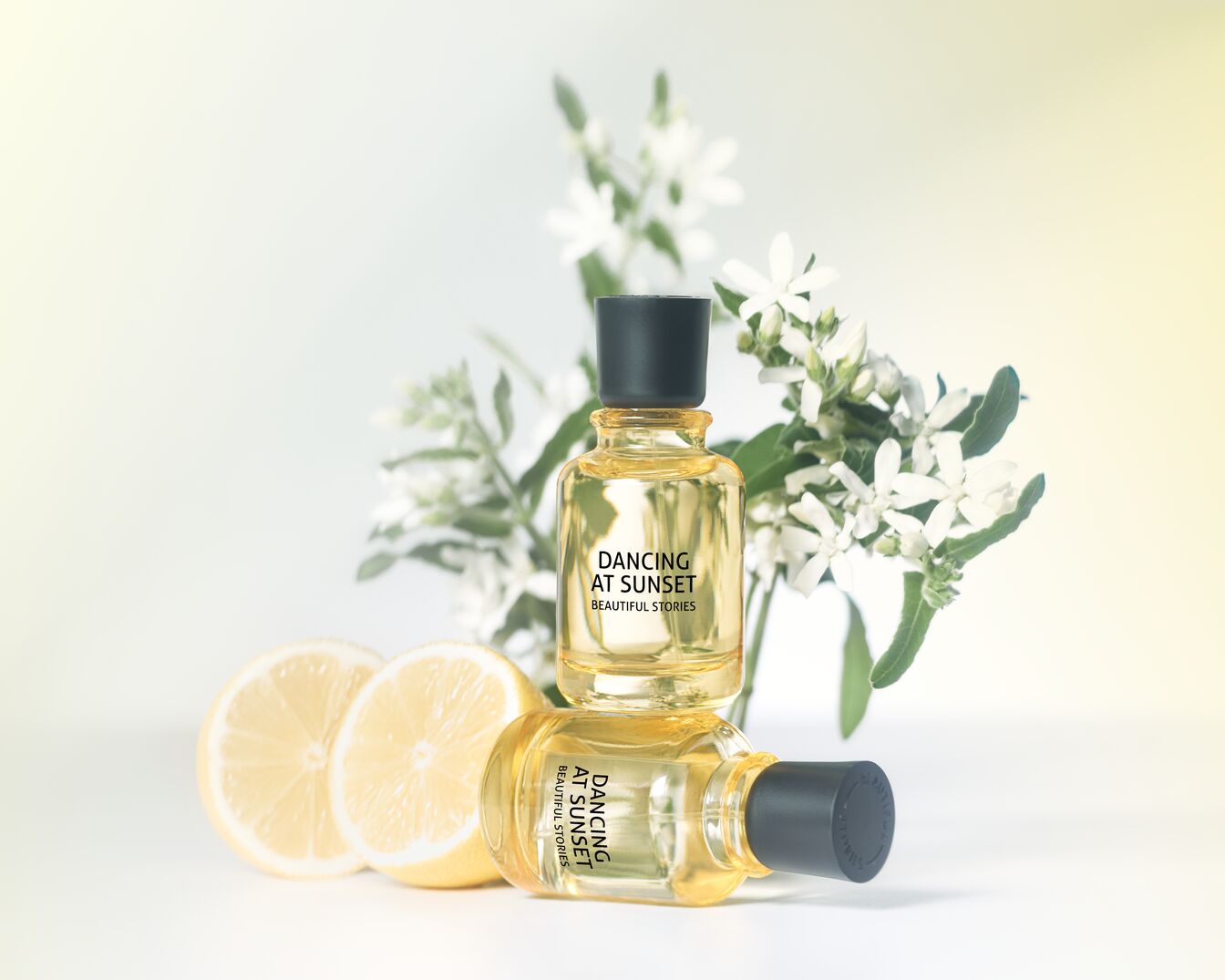 Du skaidrūs Dancing At Sunset Beautiful Stories buteliukai su juodais kamšteliais, vienas ant kito, šalia baltų gėlių ir citrinos griežinėlių.