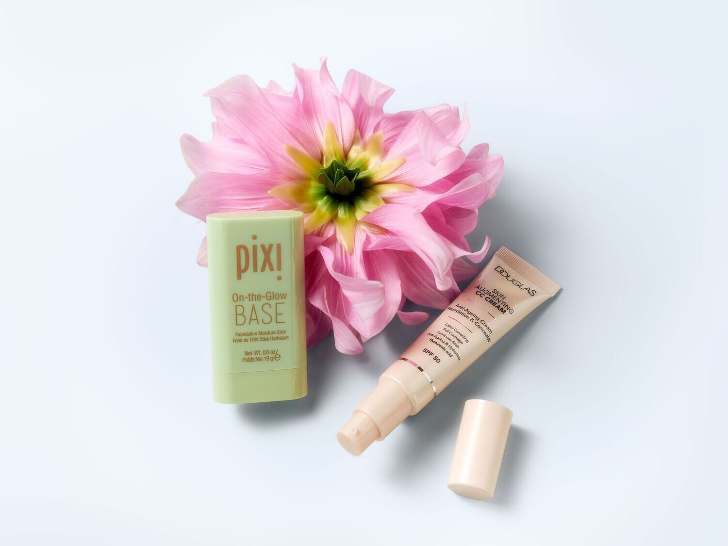 Šviesiame fone šalia rožinio žiedo guli „Pixi On-the-Glow BASE“ pagrindo lazdelė žaliame įpakavime ir „Douglas Skin Augmenting CC Cream“ kreminė pudra su SPF 50.