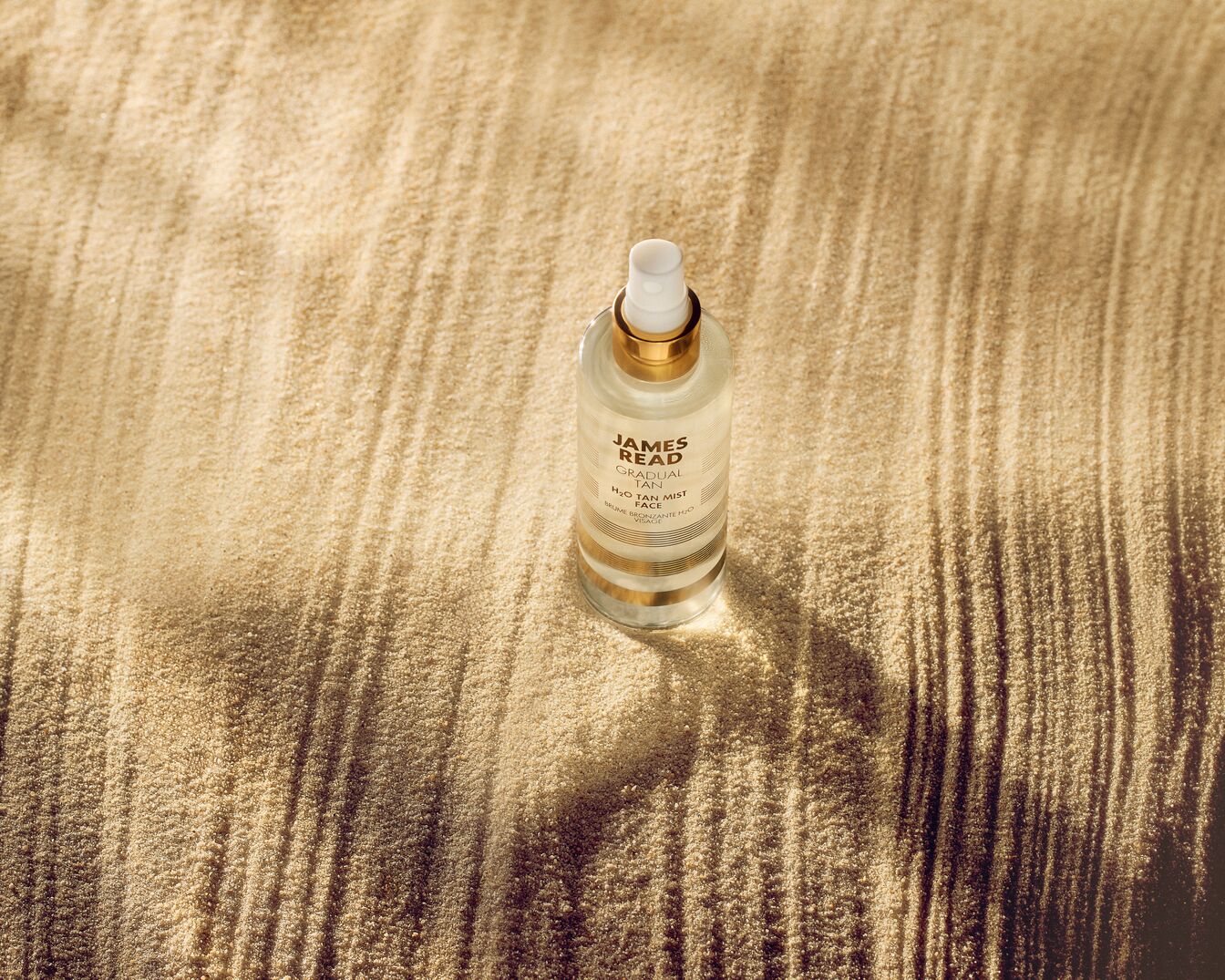 James Read Gradual Tan H2O Tan Mist Face in een heldere fles met gouden accenten, liggend op een zandstrand.