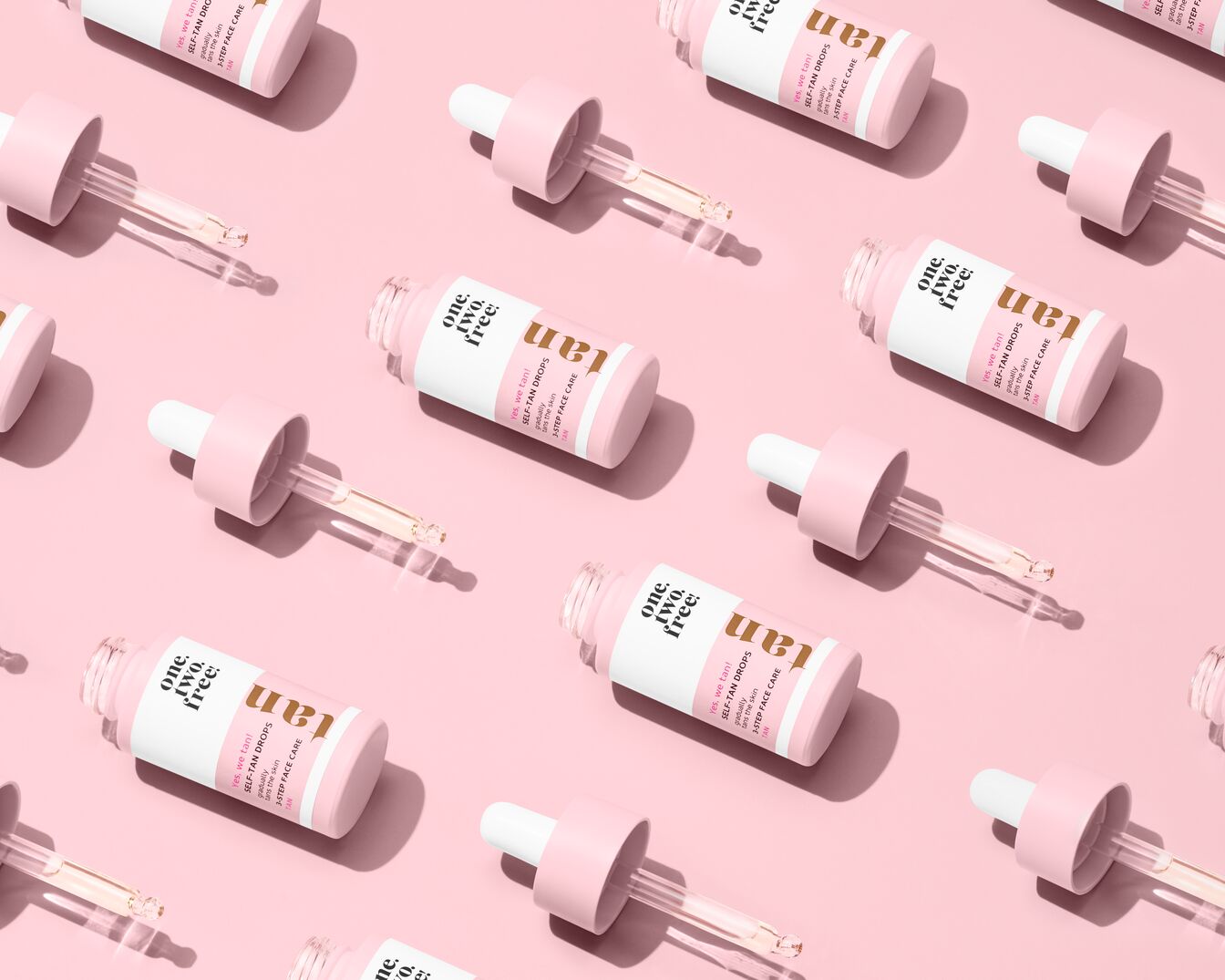 Roze achtergrond met meerdere flessen One Two Free Self-Tan Drops, die een gele vloeistof bevatten en een roze dop hebben met een glazen pipet.