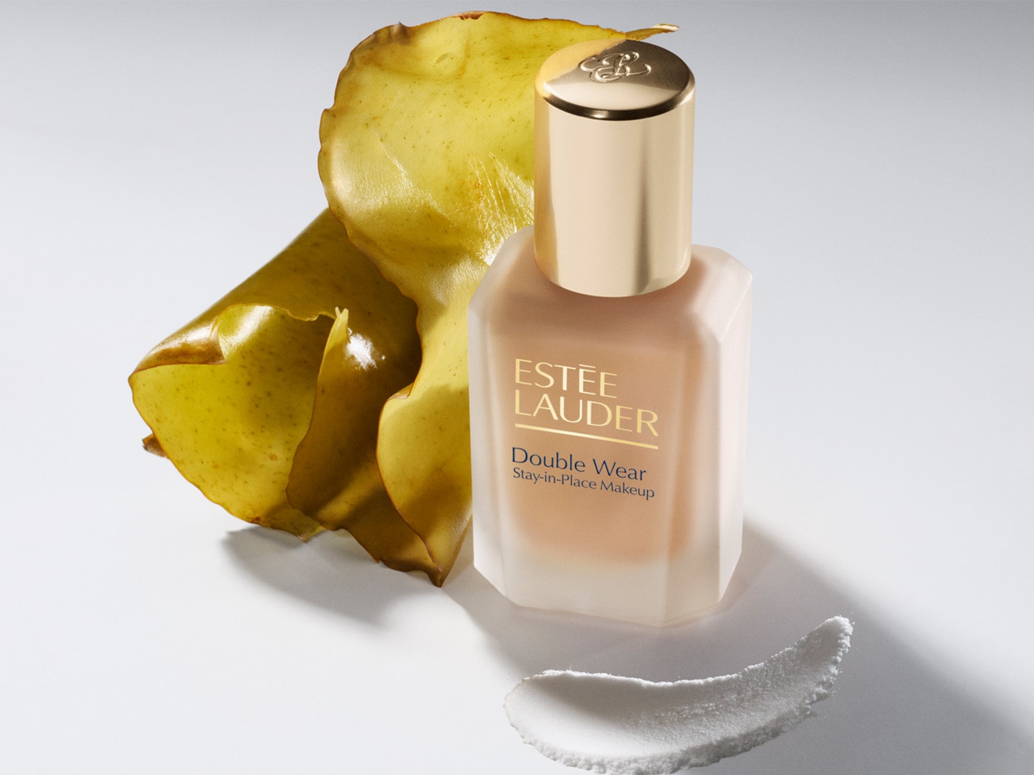 Nahaufnahme einer matten Flasche Double Wear Stay-in-Place Makeup von Estée Lauder mit goldenem Deckel, daneben ein gelbes Blatt und ein Tupfer einer weißen Creme.