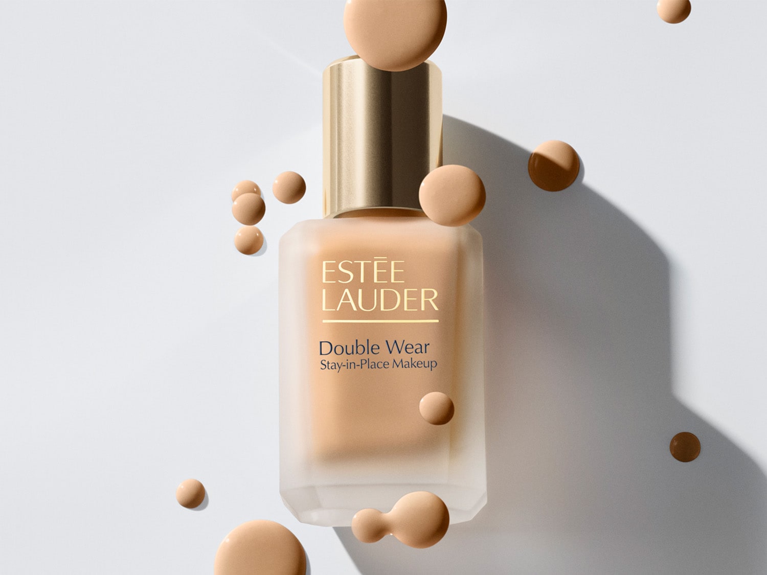 Das Double Wear Stay-in-Place Makeup von Estée Lauder in einer quadratischen, mattierten Glasflasche mit goldenem Deckel, umgeben von beigen Farbtropfen.