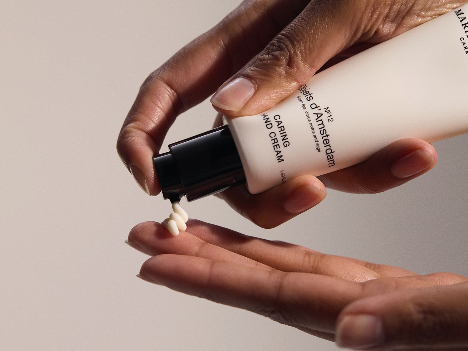 Voici une main pressant une pompe sur un tube de crème pour les mains Objets d'Amsterdam Nº12, infusée de thé vert, d'agrumes et de sauge, pour une hydratation et un soin des mains.