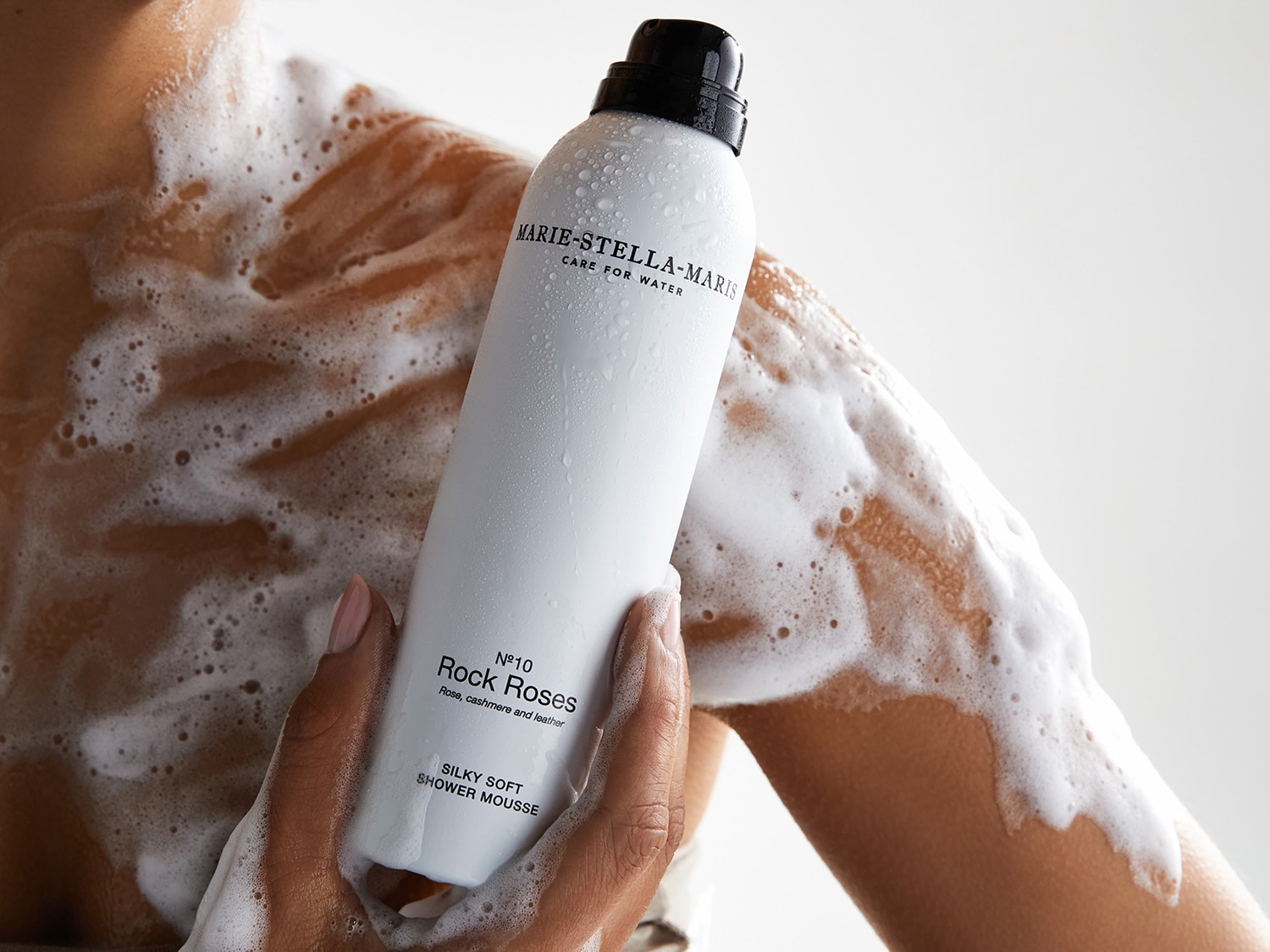 Une personne tient un flacon blanc de mousse de douche soyeuse Marie-Stella-Maris N°10 Rock Roses, avec des notes de rose, de cachemire et de cuir, tandis que de la mousse recouvre son bras et son épaule.