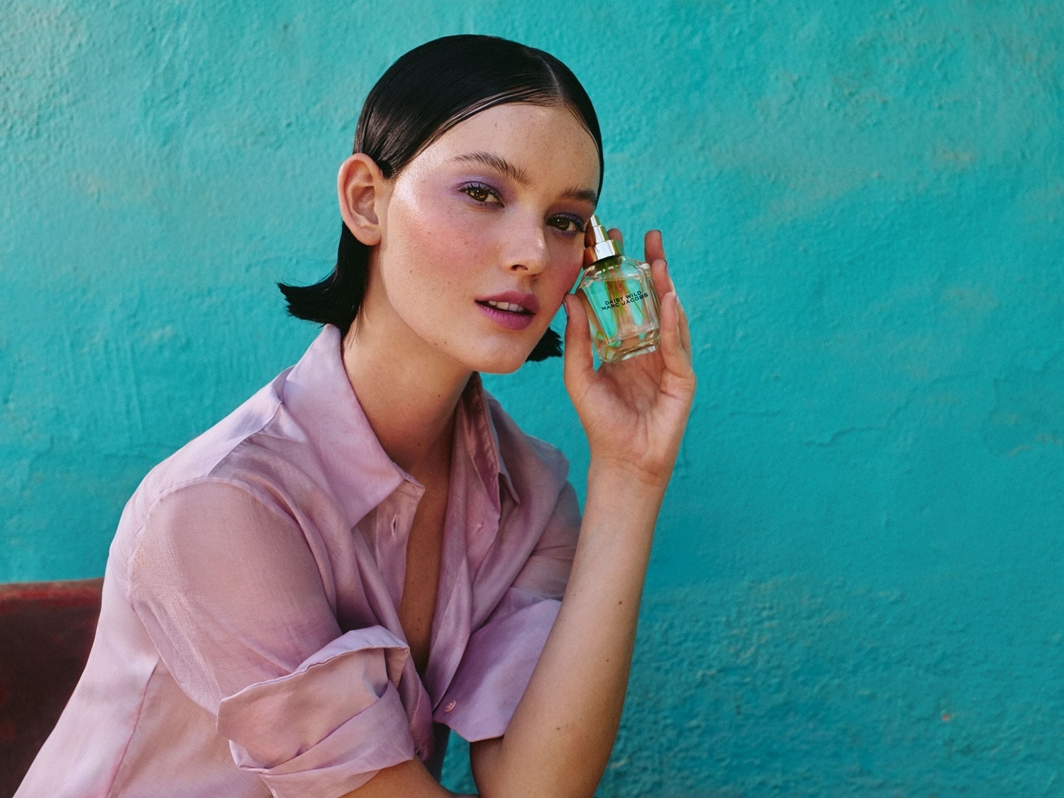 Una modella con ombretto viola e camicia rosa tiene in mano una bottiglia di profumo trasparente su uno sfondo blu brillante.