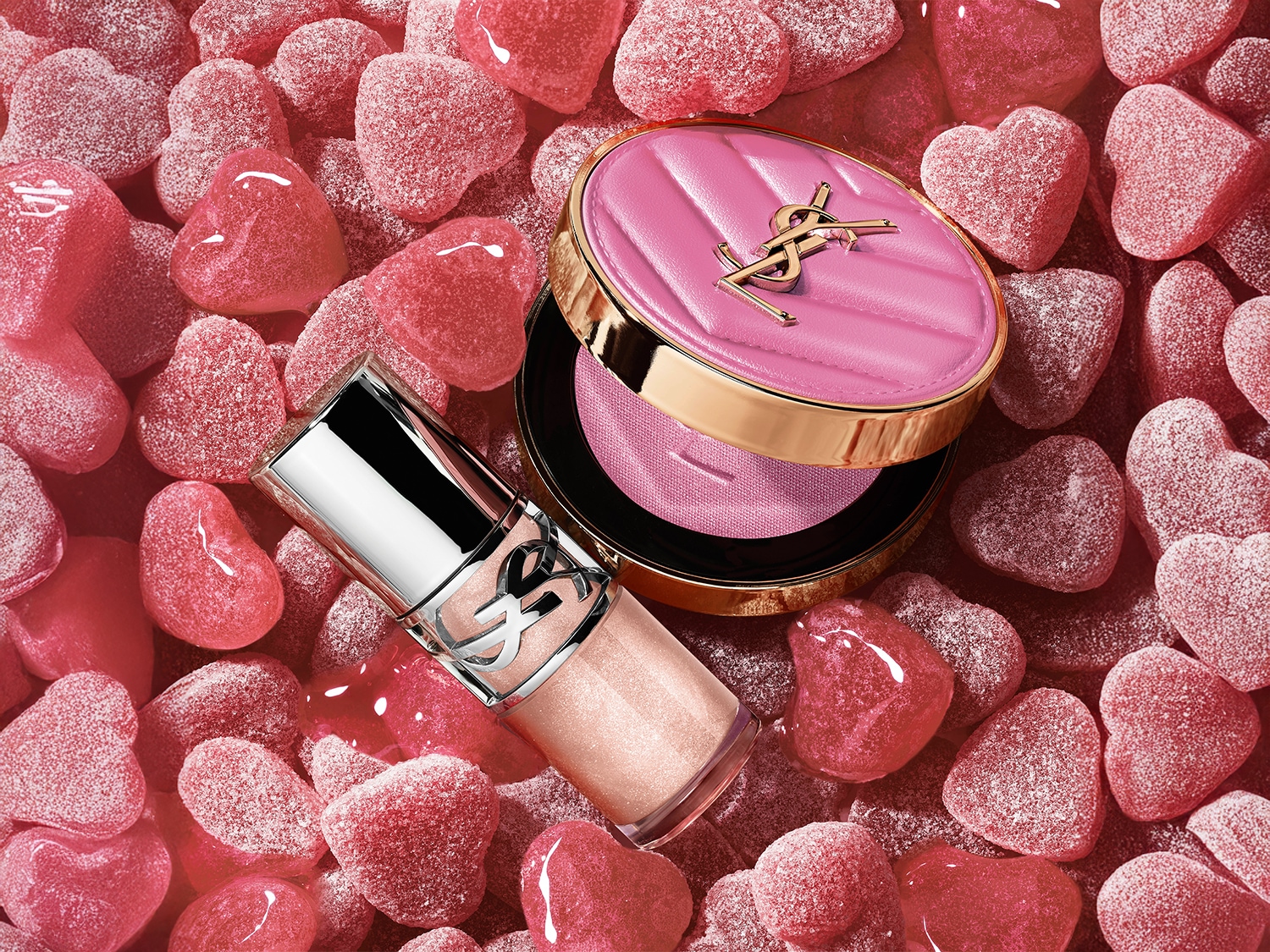 Auf einem Bett aus rosa Zuckerherzen liegen ein YSL-Highlighter mit silberner Kappe und ein Kompaktpuder mit gestepptem, rosafarbenem Etui und goldenem YSL-Logo.