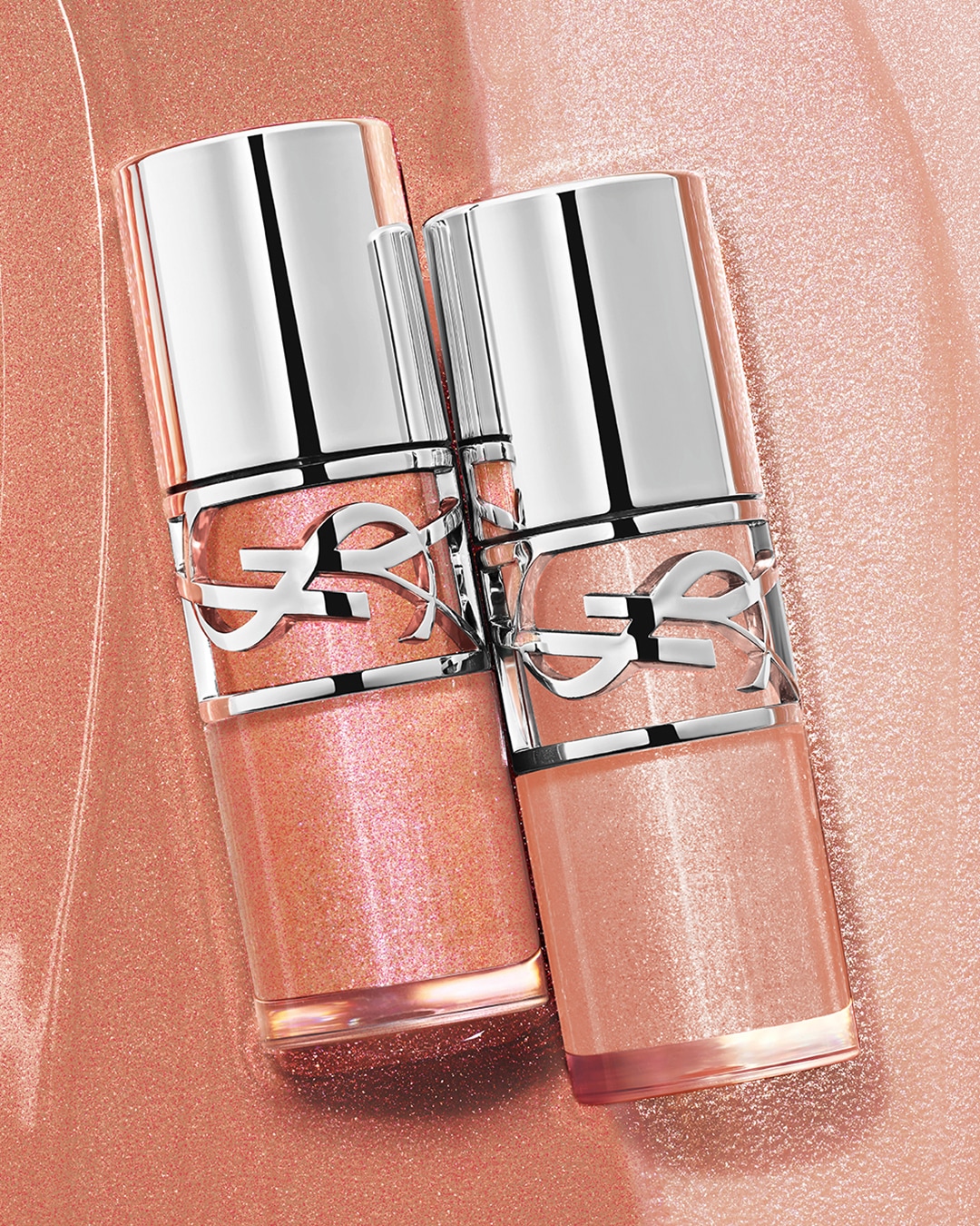 Nahaufnahme von zwei YSL-Lipgloss-Fläschchen in Pfirsichfarben mit silbernen Verschlüssen und YSL-Logo, die auf einem schimmernden, pfirsichfarbenen Untergrund liegen.