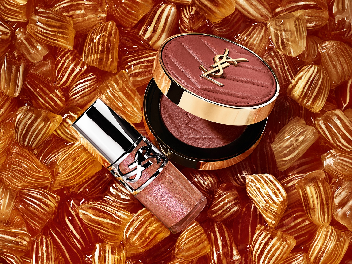 Nahaufnahme von YSL-Kosmetikprodukten, darunter ein Rouge mit goldenem Logo und ein Lipgloss, eingebettet in ein Meer von Bonbons mit Karamellwirbeln.