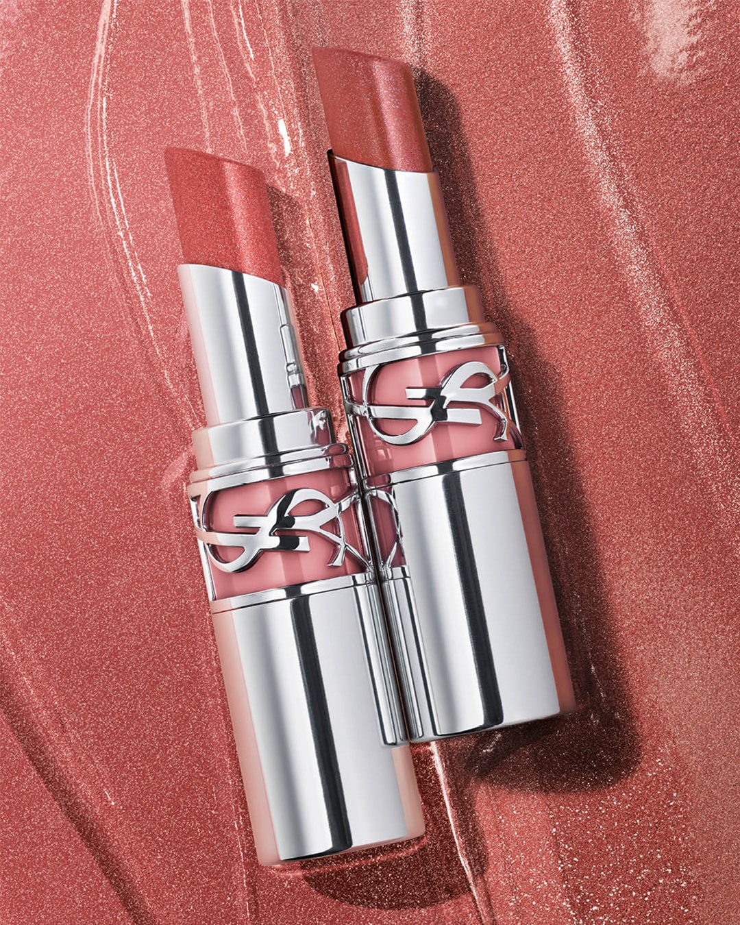 Zwei YSL Lippenstifte in silberner Verpackung mit rosa Farbtönen liegen auf einem glänzenden, rosafarbenen Untergrund.