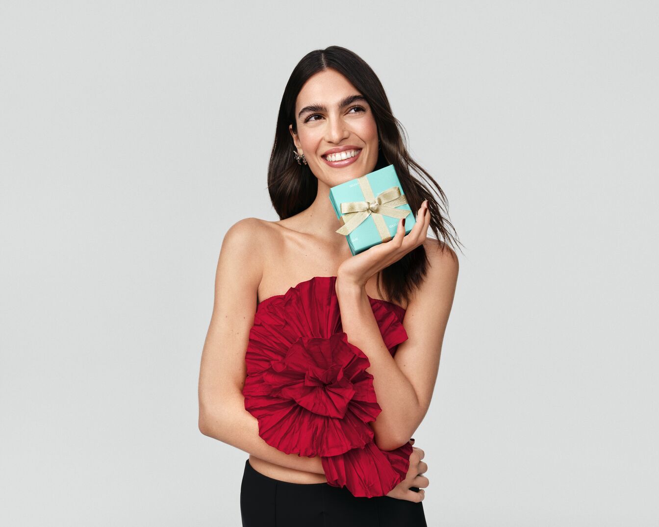 Usmívající se žena drží v ruce světle modrou dárkovou krabičku s mašlí a má na sobě červený top bez ramínek.