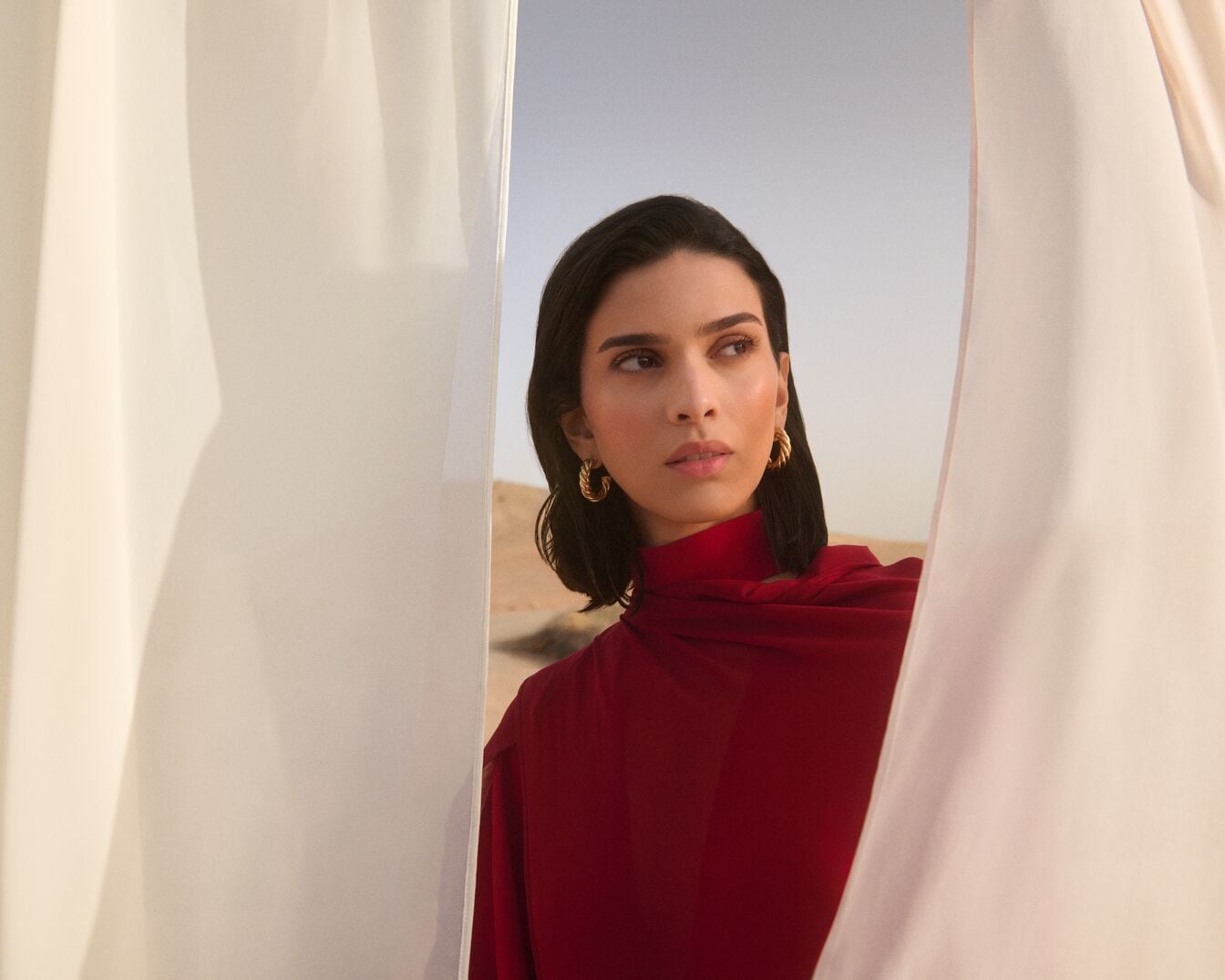 Une femme aux cheveux bruns et au chemisier rouge pose avec des boucles d'oreilles dorées, regardant à travers un rideau blanc dans un décor désertique.