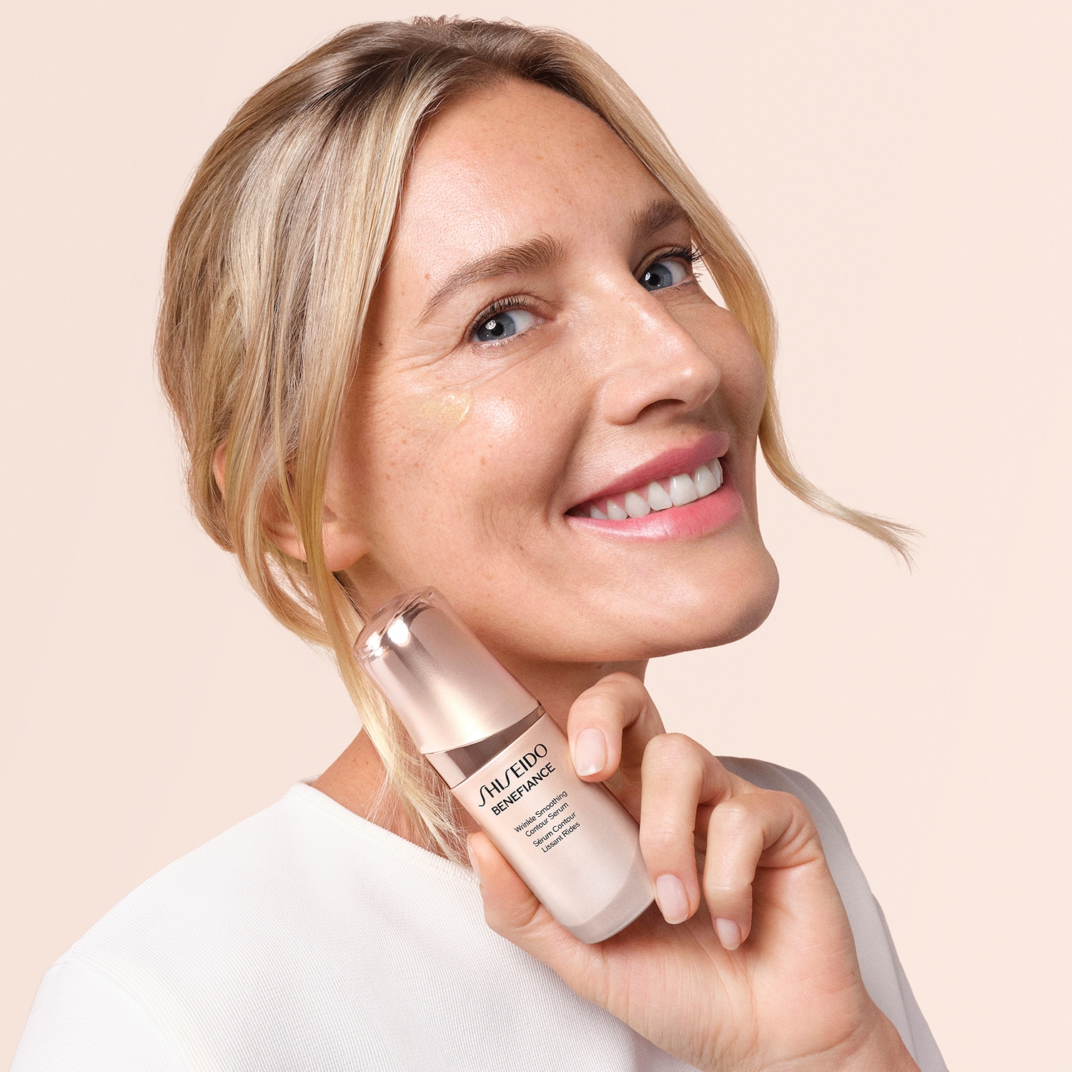 Eine blonde Frau lächelt und hält Shiseido Benefiance Wrinkle Smoothing Contour Serum in der Hand, ein Produkt zur Faltenreduzierung.