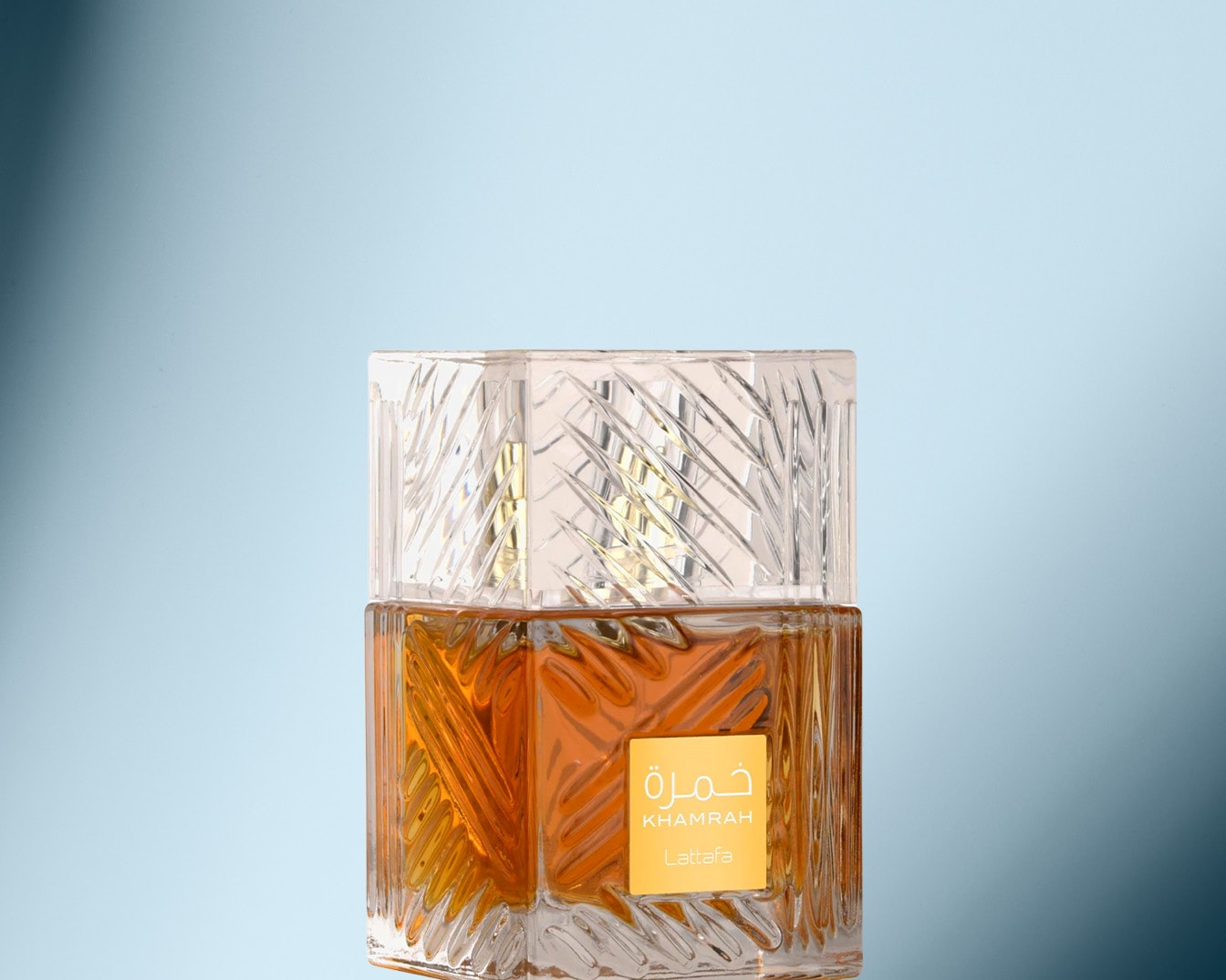 Quadratischer Khamrah-Parfümflakon von Lattafa mit durchsichtigem, strukturiertem Deckel und bernsteinfarbenem Inhalt vor hellblauem Hintergrund.