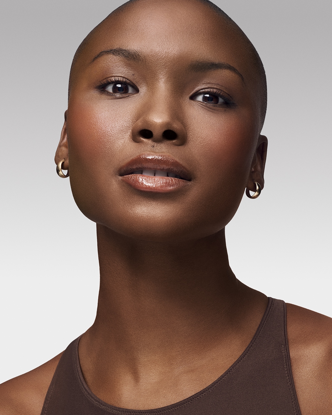 Primer plano de una mujer de piel oscura con maquillaje natural, que resalta sus ojos marrones y labios brillantes, con aros dorados y una camiseta marrón.