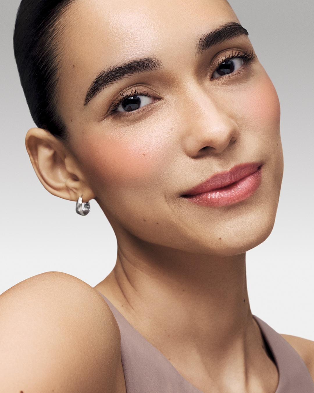 Primer plano de una mujer con maquillaje natural, que incluye rubor rosa en las mejillas y brillo labial rosa brillante, complementado con un pendiente plateado.