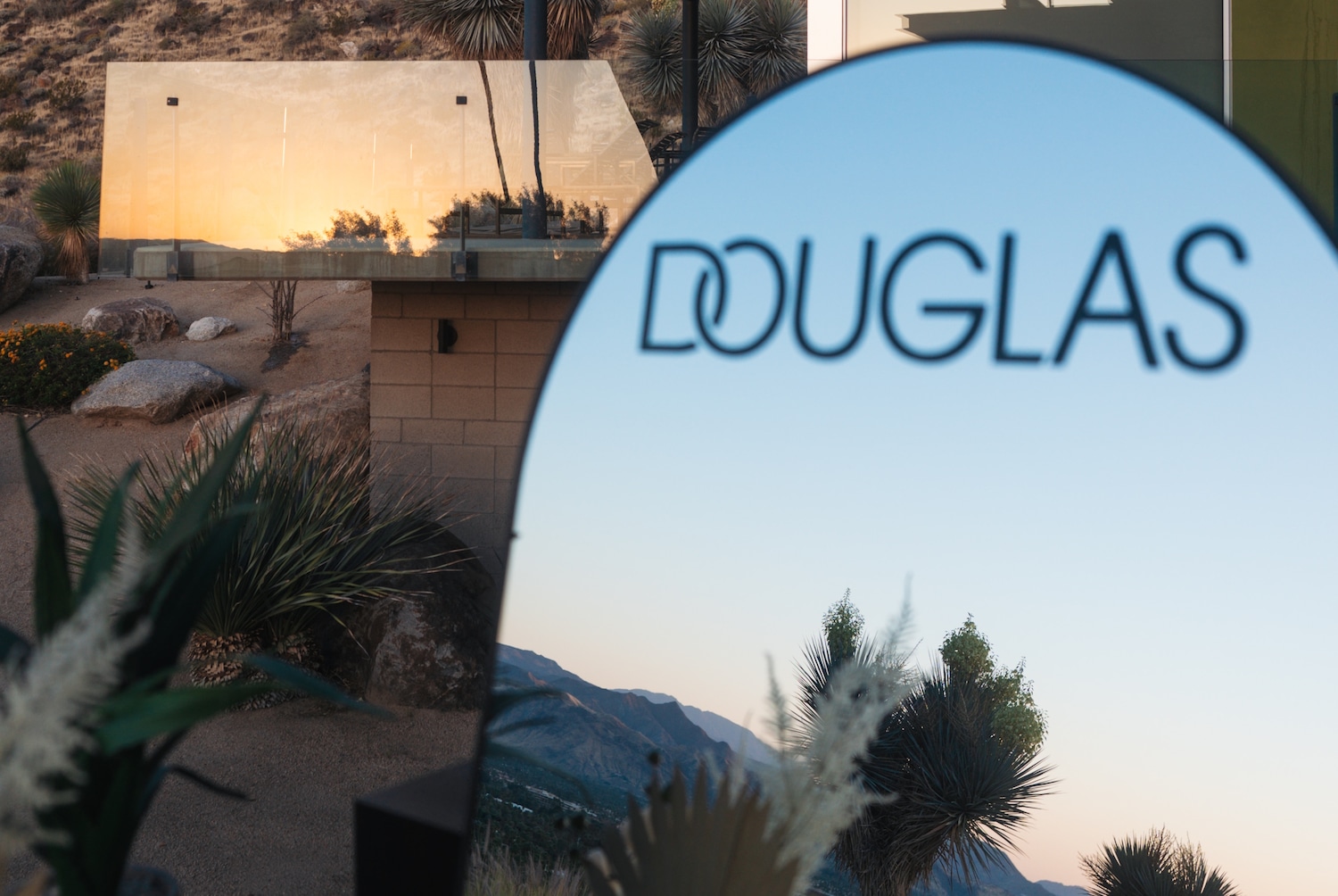 Das Douglas-Logo vor einer Wüstenlandschaft mit Bergen im Hintergrund, die sich im Spiegelglas eines Gebäudes widerspiegeln.