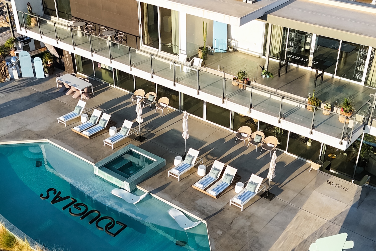 Vogelperspektive auf ein modernes zweistöckiges Gebäude mit einem Pool, auf dem der Name DOUGLAS in großen Buchstaben steht, umgeben von Liegestühlen und Sonnenschirmen.