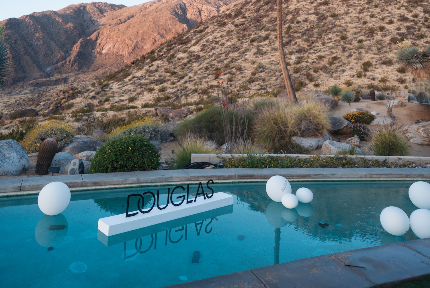 Ein Pool mit dem Schriftzug DOUGLAS auf einem weißen Brett, umgeben von weißen Ballons, vor einer Wüstenlandschaft mit Bergen im Hintergrund.