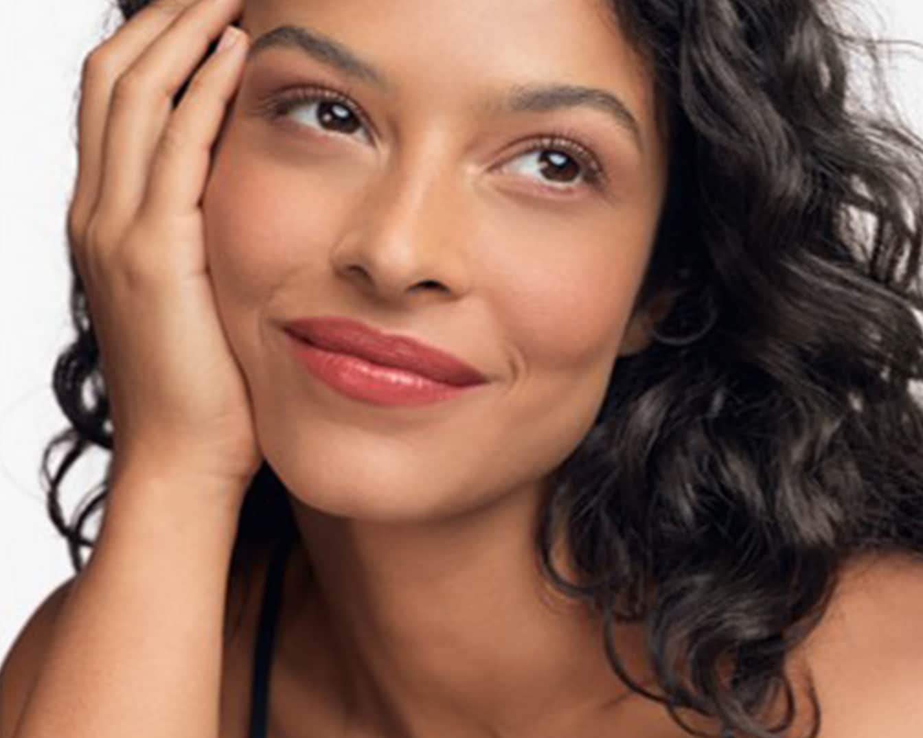 Primo piano di una donna con capelli scuri e ondulati, pelle abbronzata e labbra color corallo, che appoggia il viso sulla mano con uno sguardo sognante.