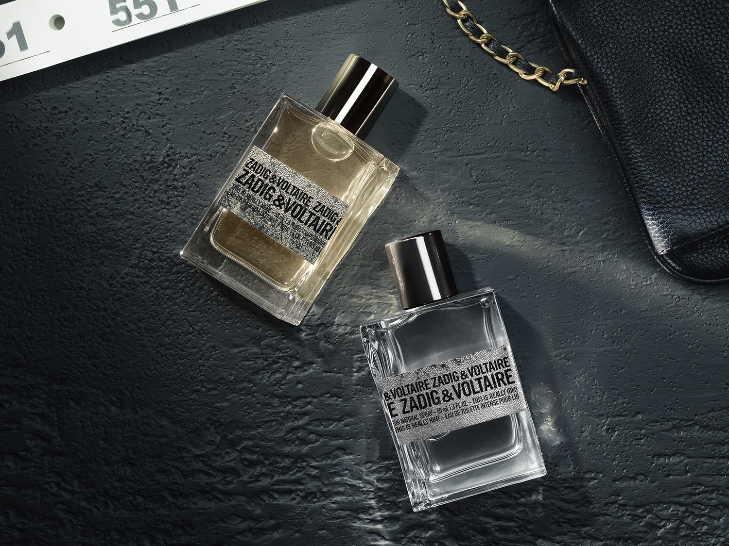 Zwei ZADIG&VOLTAIRE Düfte aus der This Is Really! Linie liegen auf einer dunklen, strukturierten Oberfläche. Die klaren Glasflakons mit metallischen Kappen wirken urban, modern und markant. Eine schwarze Tasche mit Kettendetail ergänzt das stilvolle, edgy Setting.