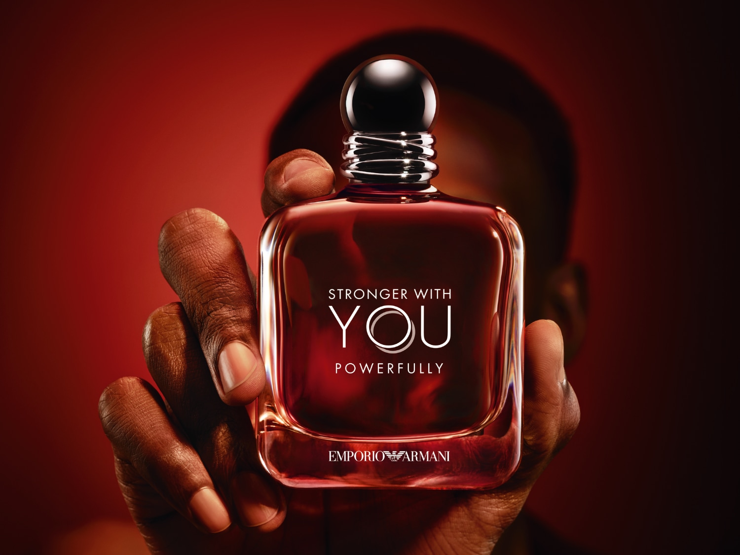 Detailní záběr na flakon parfému Emporio Armani Stronger With You, který drží v ruce muž s tmavou pletí na červeném pozadí.
