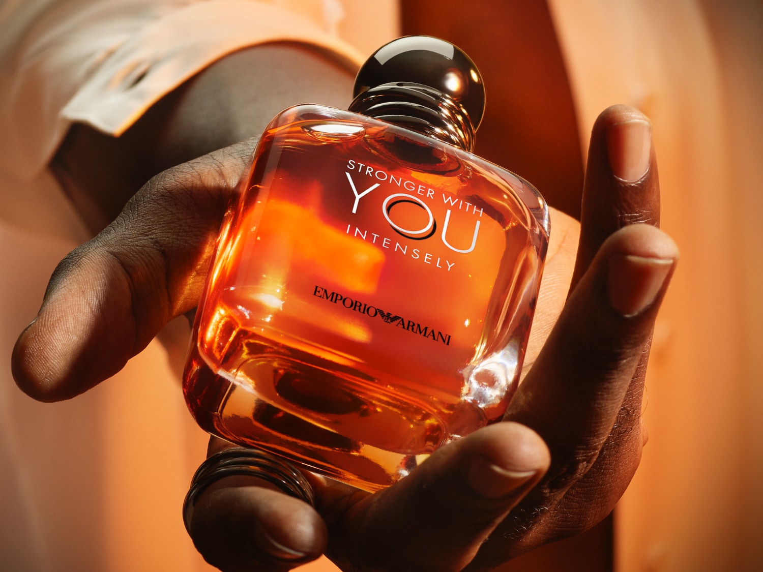 Detailný záber na flakón pánskeho parfumu Emporio Armani Stronger With You Intensely s oranžovým nádychom, ktorý drží v ruke muž s prsteňmi.