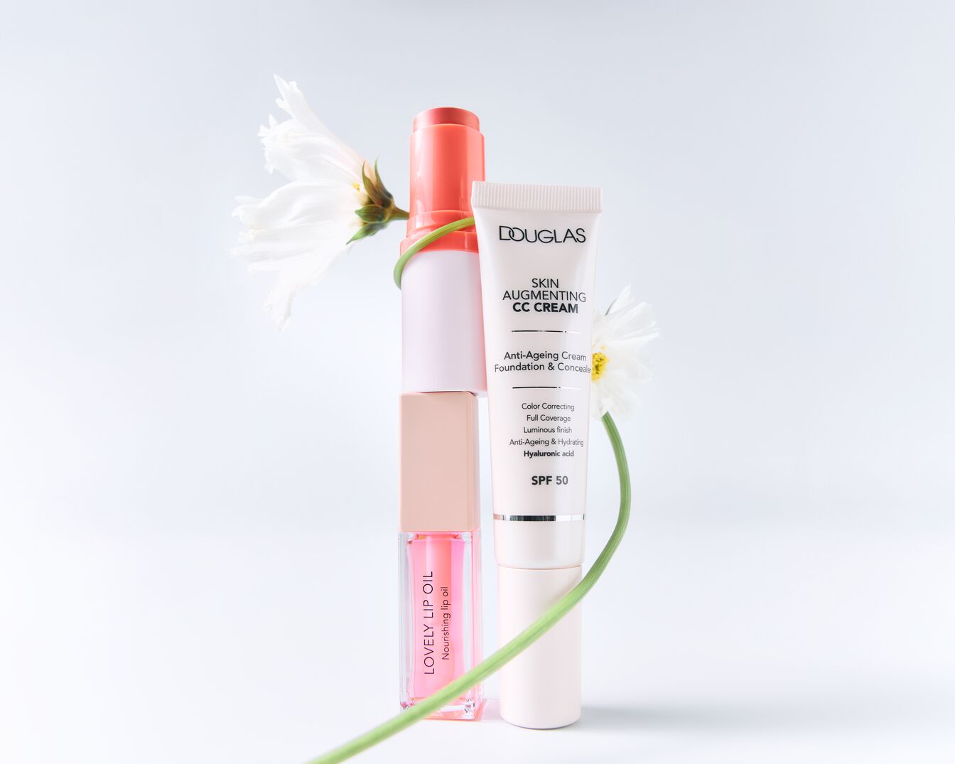 Pleťový olej Lovely Lip Oil, CC krém Douglas Skin Augmenting a rtěnka jsou zobrazeny na bílém pozadí, doplněné bílou květinou.