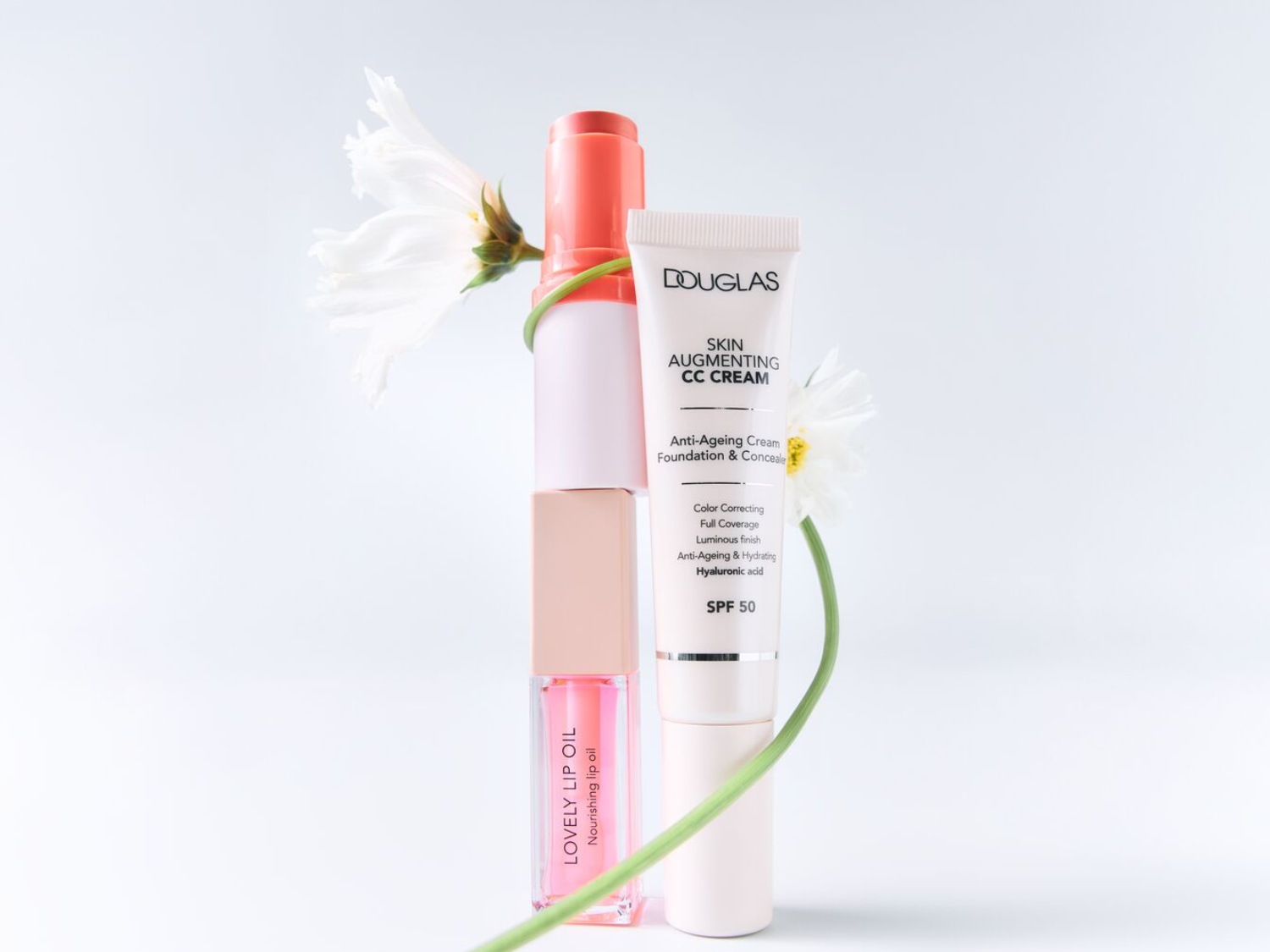 Na obrázku je zobrazený rad kozmetických produktov, vrátane Lovely Lip Oil, Douglas Skin Augmenting CC Cream s SPF 50, anti-ageingovým krémom, make-upom a korektorom, všetko doplnené bielymi kvetmi.