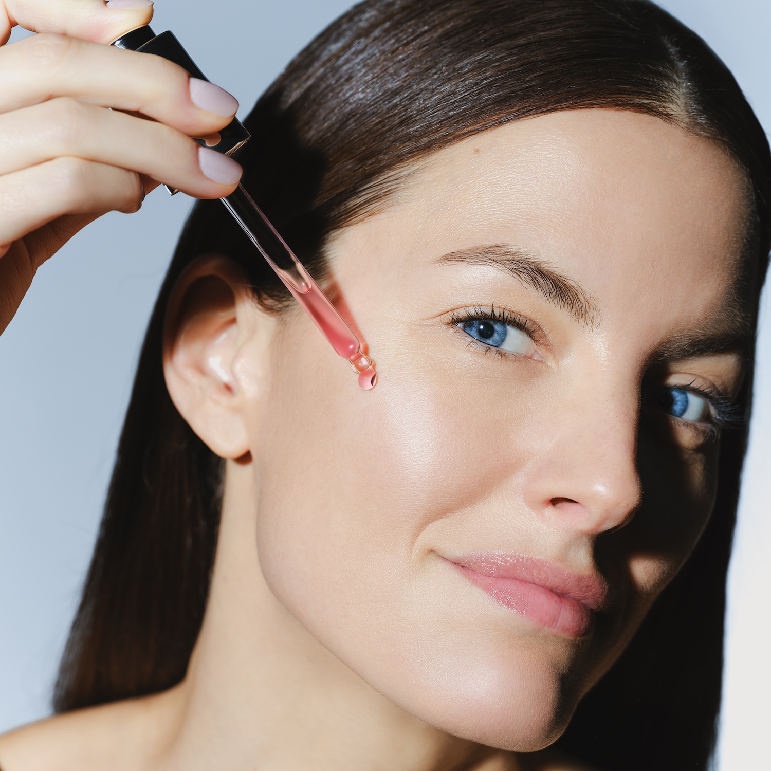 Une femme applique un sérum rose sur sa joue avec une pipette, mettant en valeur une peau éclatante et des yeux bleus lumineux.