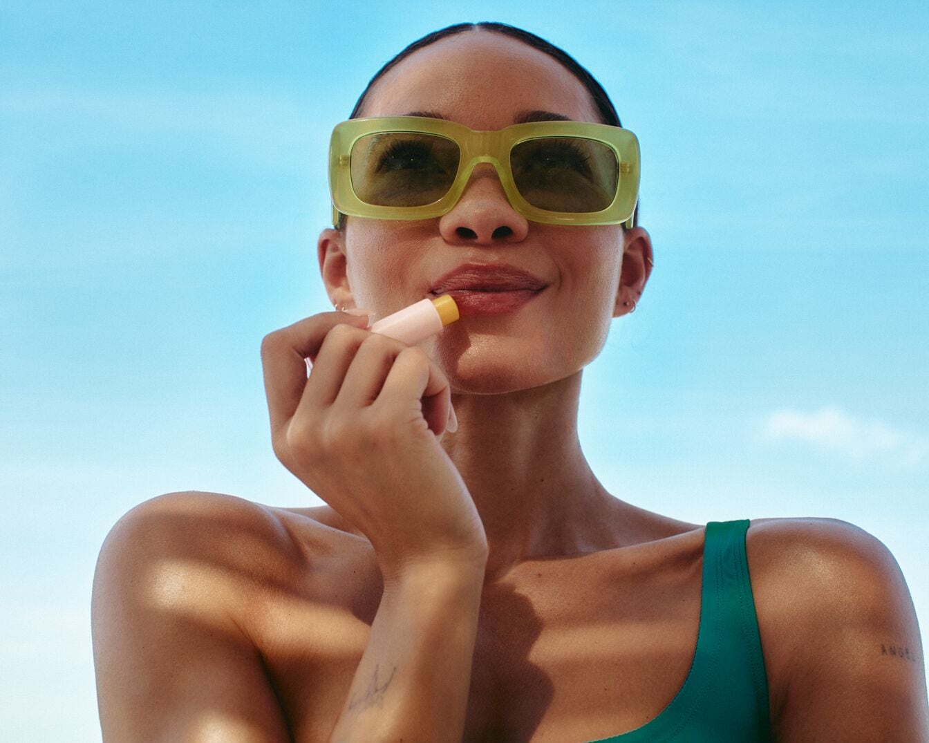 Sotto il cielo azzurro, una donna con occhiali da sole verde chiaro applica un balsamo labbra color pesca, esaltando la naturale bellezza delle sue labbra.