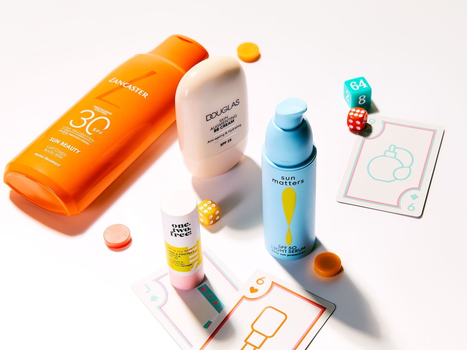 Nuotraukoje matomi įvairūs apsaugos nuo saulės produktai, įskaitant „Lancaster“ losjoną su SPF 30, „Douglas BB“ kremą su SPF 25 ir „Sun Matters“ serumą su SPF 50, išdėlioti ant balto paviršiaus su žaidimo elementais.