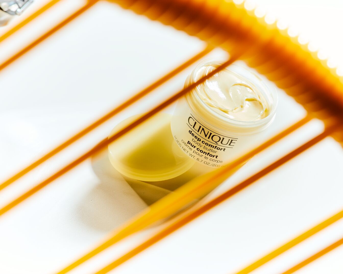 Unt de corp Clinique Deep Comfort într-un borcan deschis, cu o textură cremoasă, așezat pe un fundal alb cu linii portocalii.