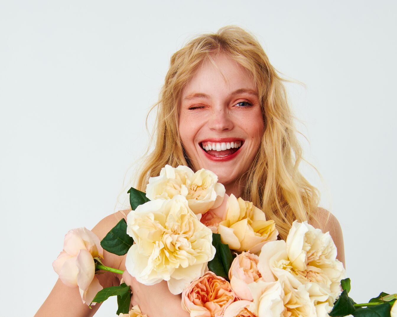 Blonďatá žena s úsměvem a mrknutím drží kytici žlutých a broskvových květů.