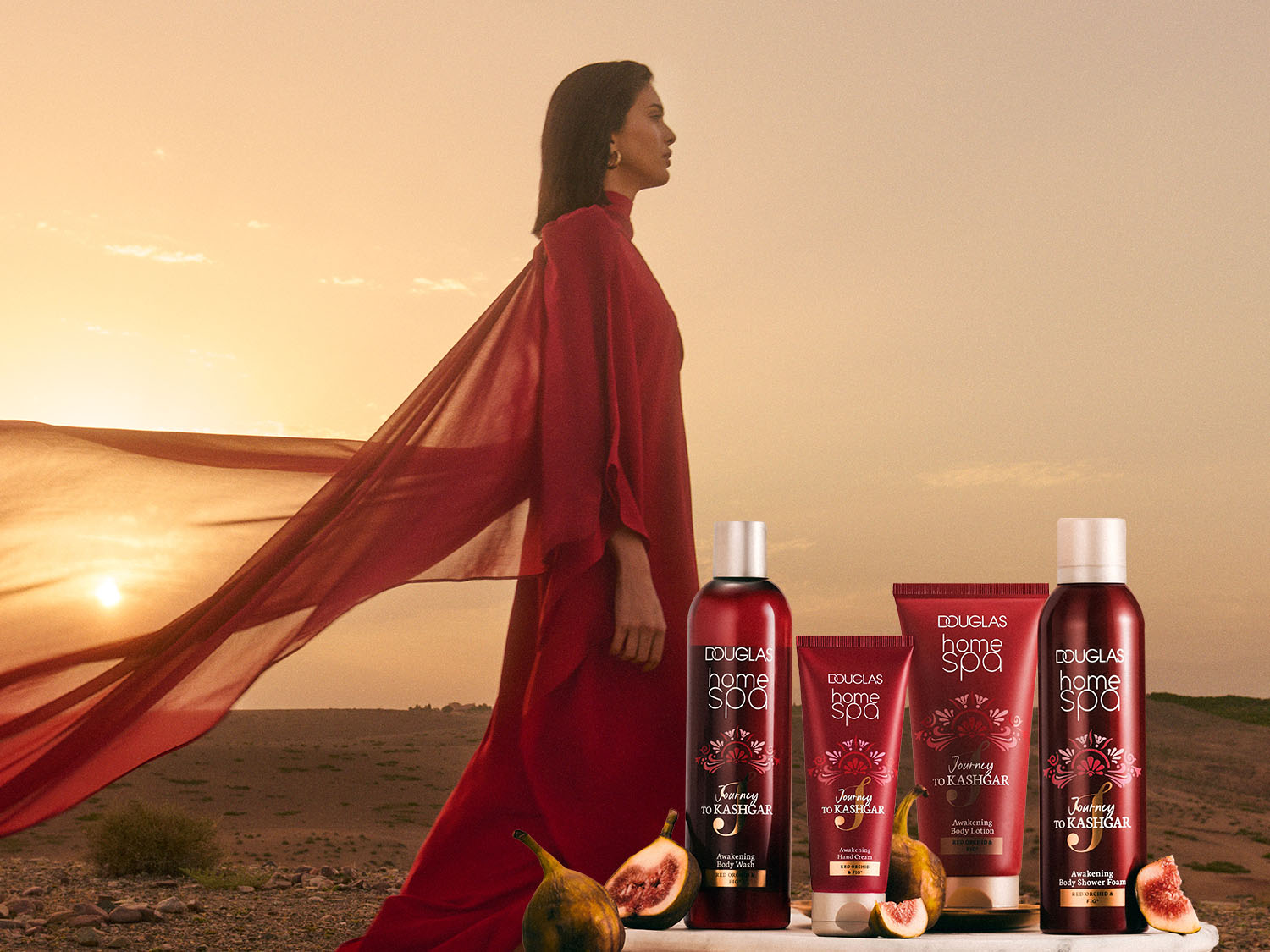 Eine Frau in rotem Kleid steht in der Wüste neben Douglas Home Spa Produkten, darunter Body Wash, Handcreme, Bodylotion und Duschschaum mit dem Duft von roter Orchidee und Feige.