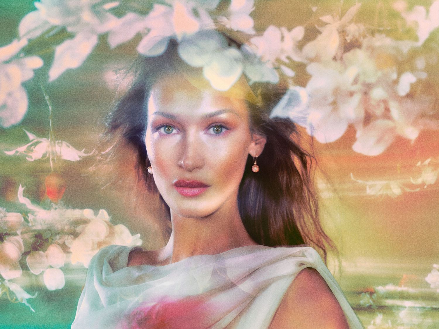Portret van Bella Hadid met los, donkerbruin haar, groene ogen en roze lippen, gehuld in een witte doek, omringd door witte bloemen.