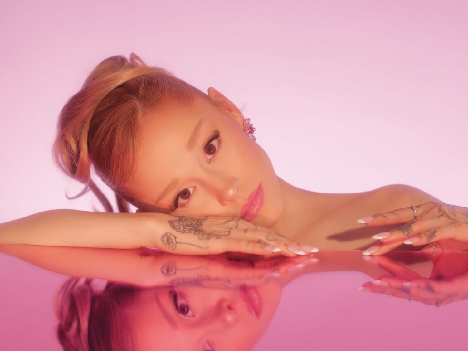 Een close-up van Ariana Grande met een roze achtergrond, haar roze haar in een knot, ze rust op haar handen met een zachte roze lipgloss.