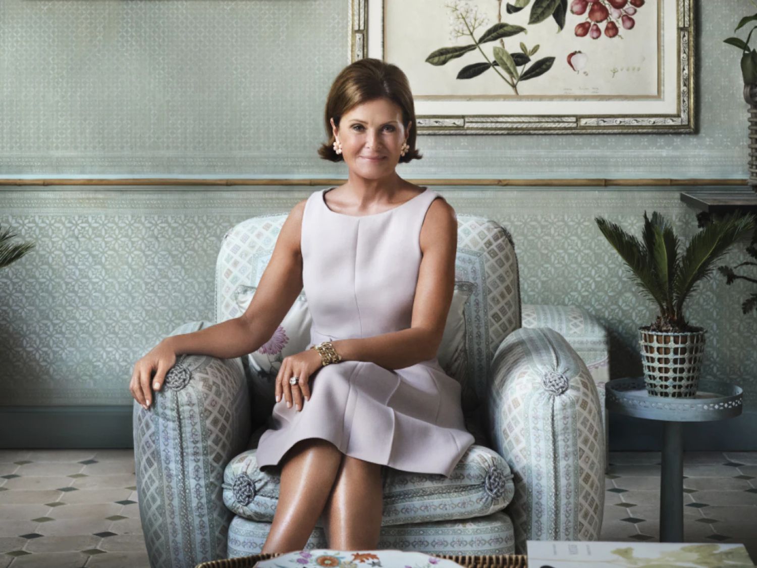Een elegante vrouw met kort bruin haar zit in een sierlijke fauteuil, gekleed in een lichtroze jurk, met een schilderij van botanische illustraties op de achtergrond.