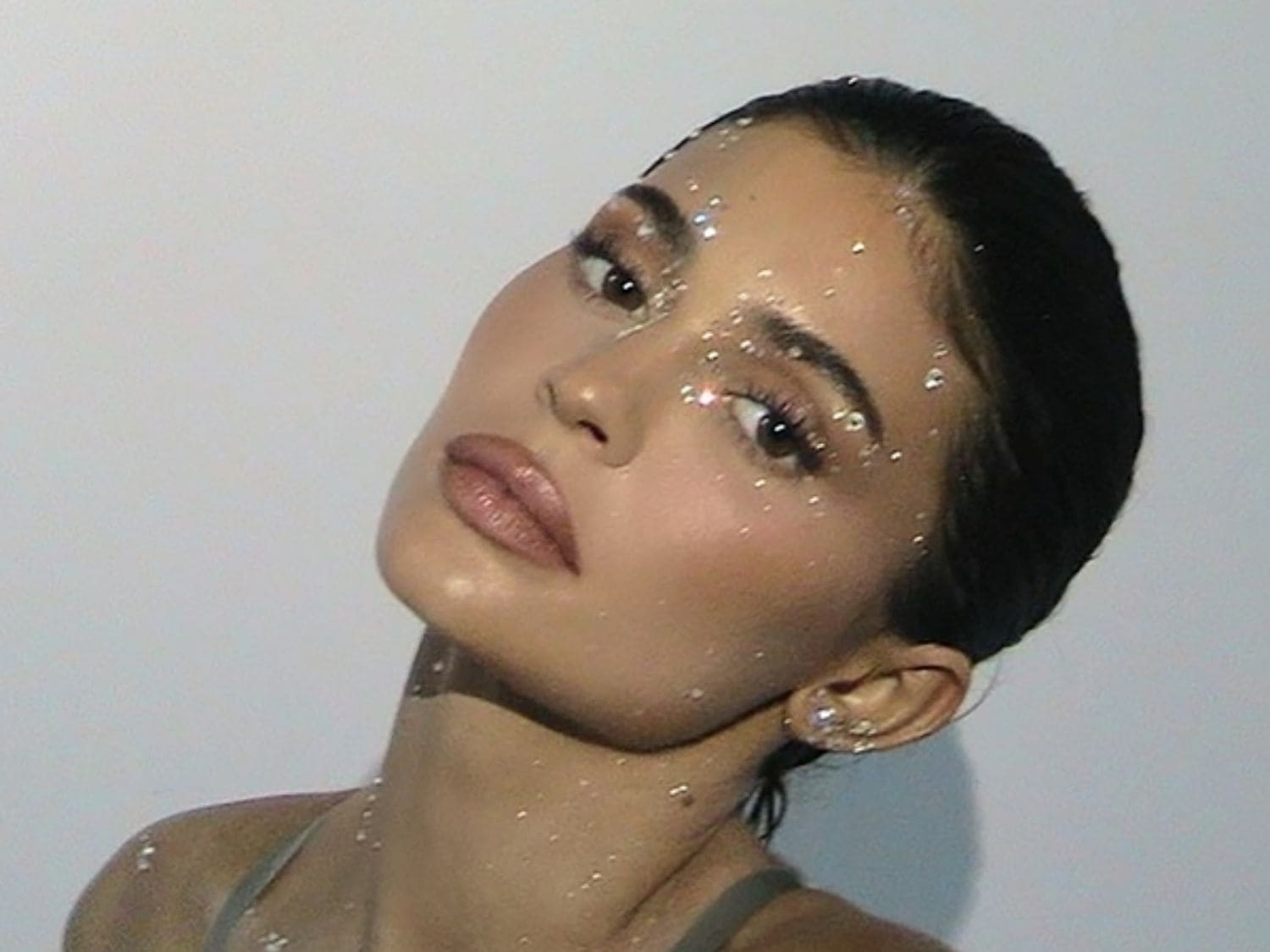 Een vrouw met glitters op haar gezicht en lichaam, haar make-up is nude en haar haar is strak naar achteren gekamd.