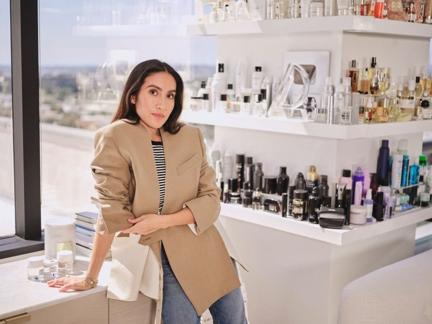 Jen Atkin met een beige blazer staat in een kamer vol schoonheidsproducten, klaar om haar favorieten te delen.