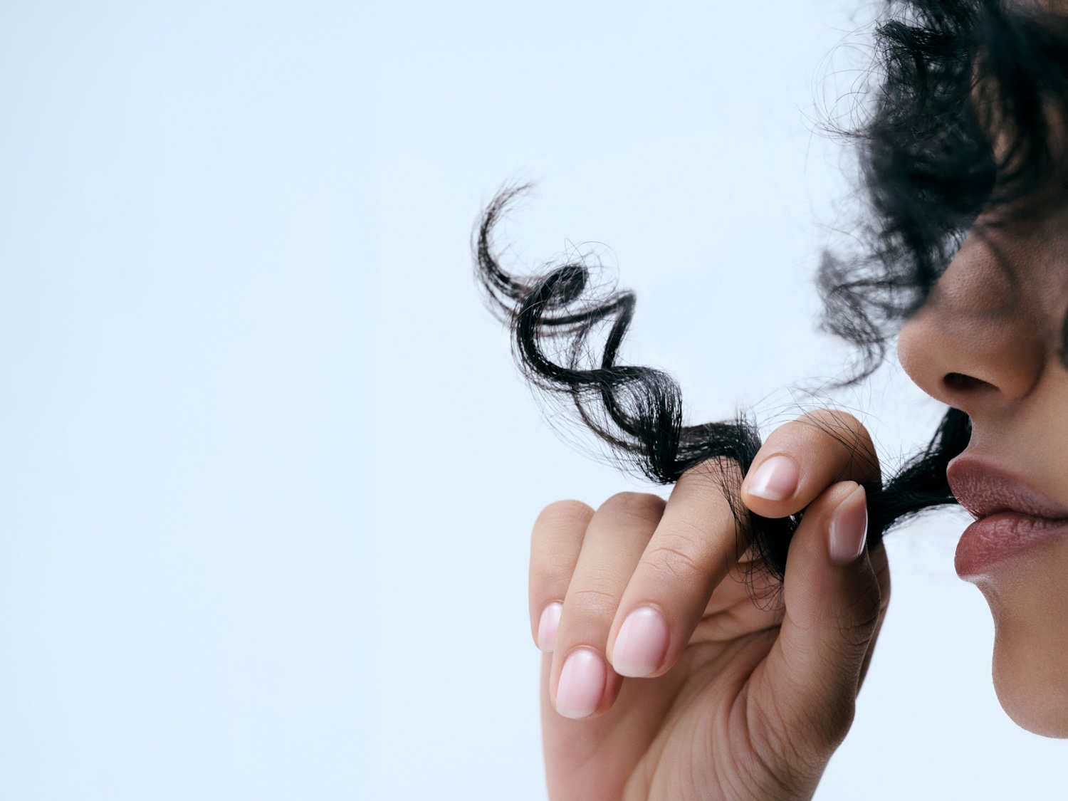 Una donna con la pelle scura e le labbra rosa chiaro tiene in mano una ciocca di capelli ricci neri, con le unghie curate e smaltate di rosa chiaro.