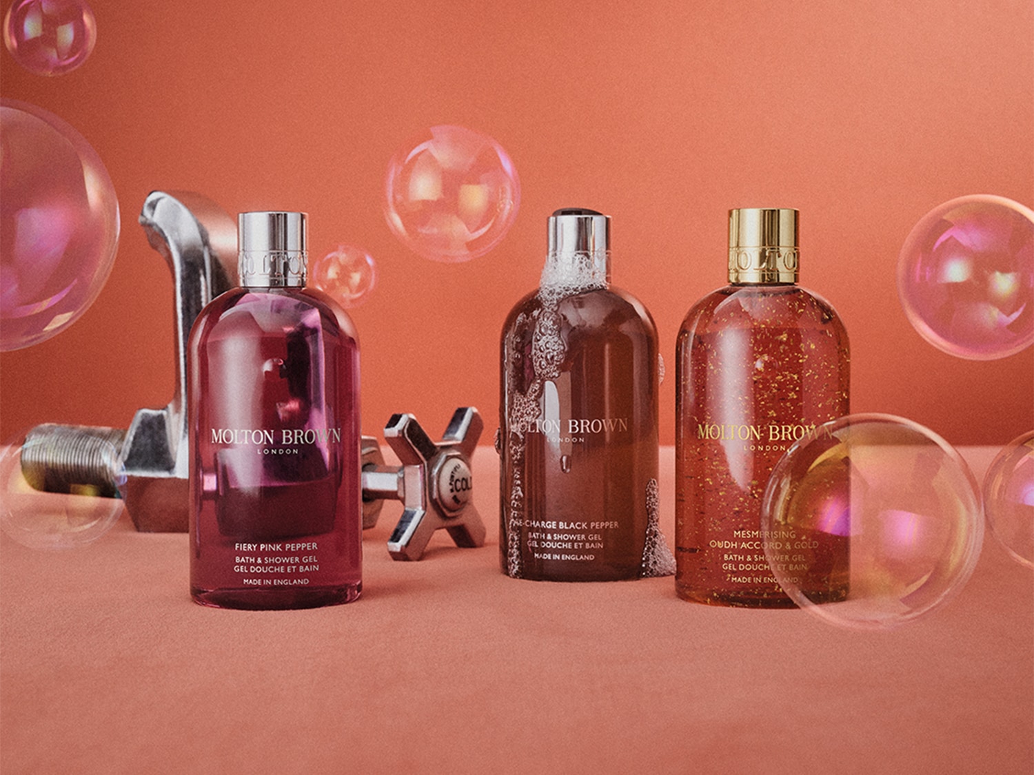 Drei transparente Molton Brown London Flaschen mit silbernen, goldenen und silbernen Verschlüssen, die Duschgel in Rosa-, Braun- und Goldtönen enthalten.