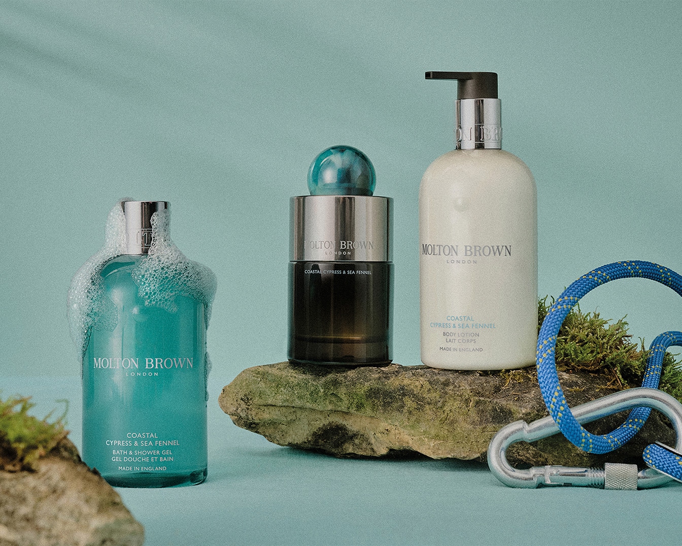 Drei Molton Brown London Produkte der Coastal Cypress & Sea Fennel Linie, darunter ein Badeschaum, ein Eau de Parfum und eine Bodylotion, auf einem Felsen arrangiert.