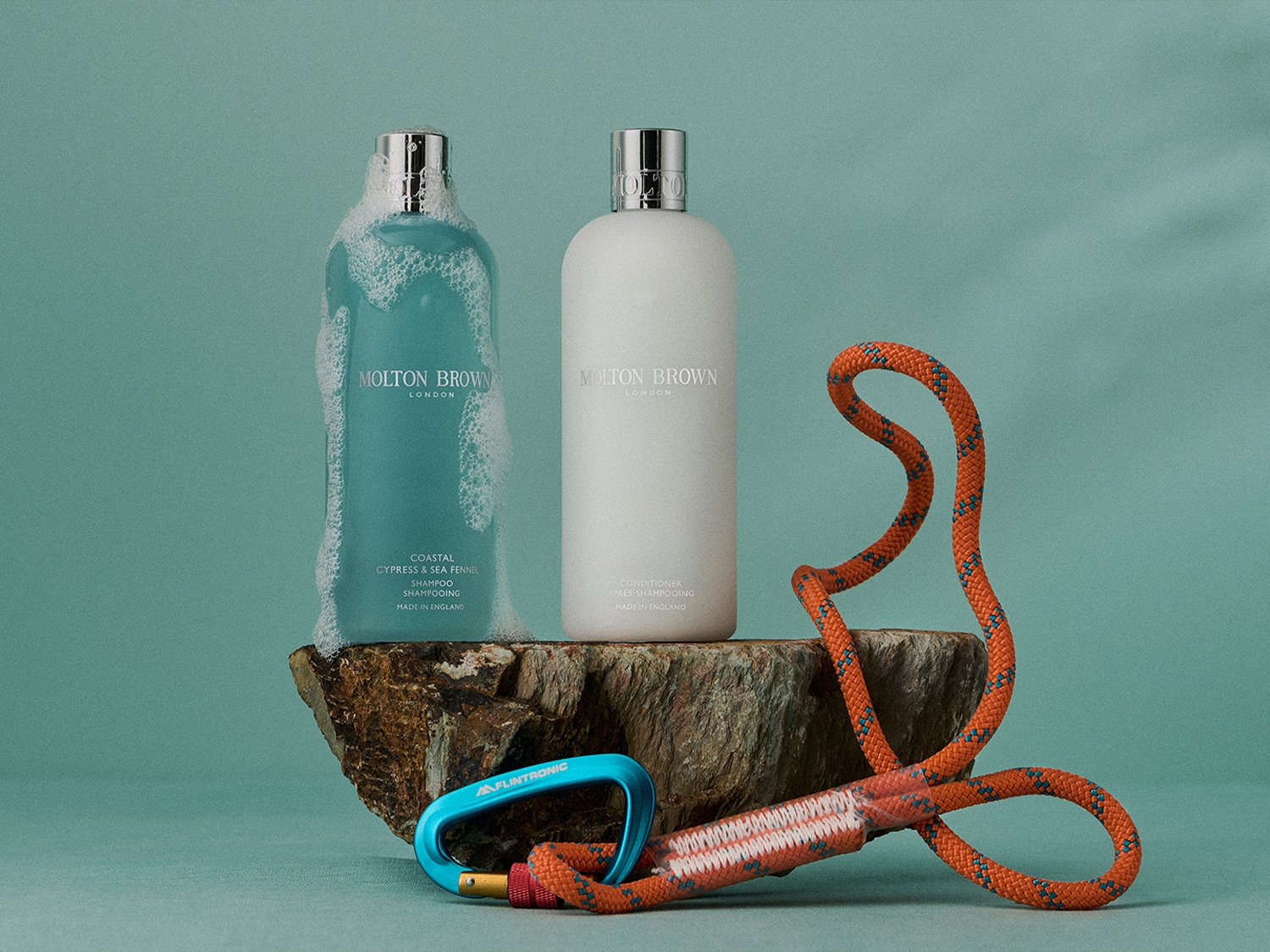 Zwei Flaschen Molton Brown London, eine türkisfarbene Coastal Cypress & Sea Fennel Shampoo-Flasche mit Schaum und eine weiße Conditioner-Flasche, stehen auf einem Felsen, dekoriert mit einem orangefarbenen Seil und Karabiner.