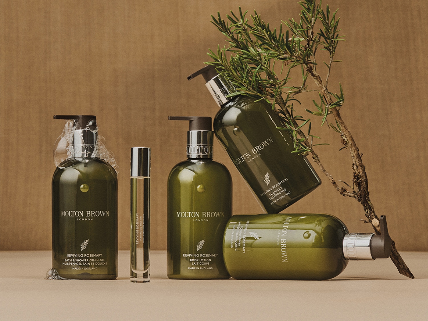 Eine Kollektion von Molton Brown Reviving Rosemary Produkten, darunter Duschgel, Körperlotion, Parfümöl und Shampoo, arrangiert mit einem Zweig Rosmarin.