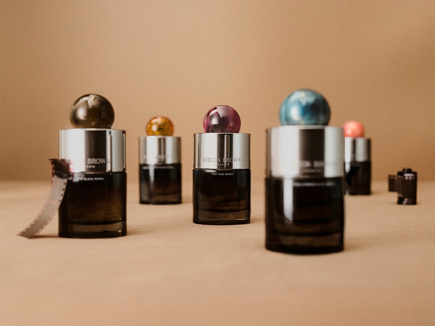 Eine Nahaufnahme von Molton Brown Parfümflaschen mit silbernen Verschlüssen und farbigen Kugeln auf einem beigen Hintergrund, die eine elegante und moderne Ästhetik präsentieren.