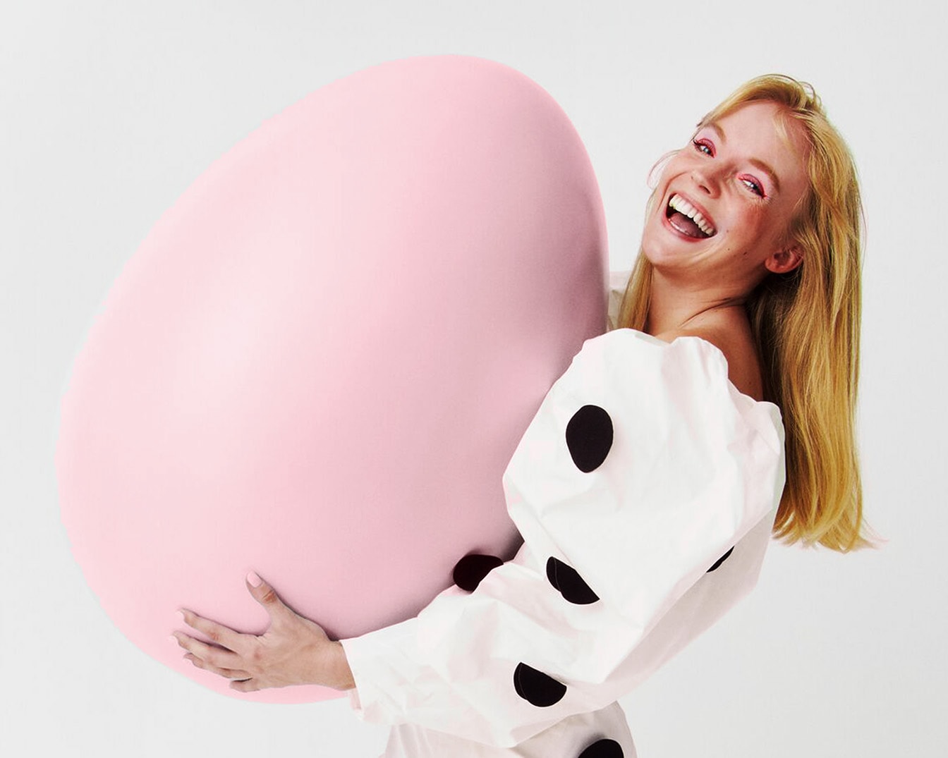 Une femme blonde souriante porte un grand œuf rose, vêtue d'une blouse blanche à pois noirs, pour une ambiance festive et joyeuse.