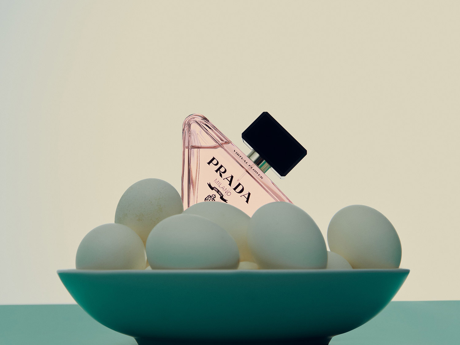 Un flacon de parfum Prada rose repose sur un bol turquoise rempli d'œufs blancs, créant un contraste visuel intrigant et inattendu.