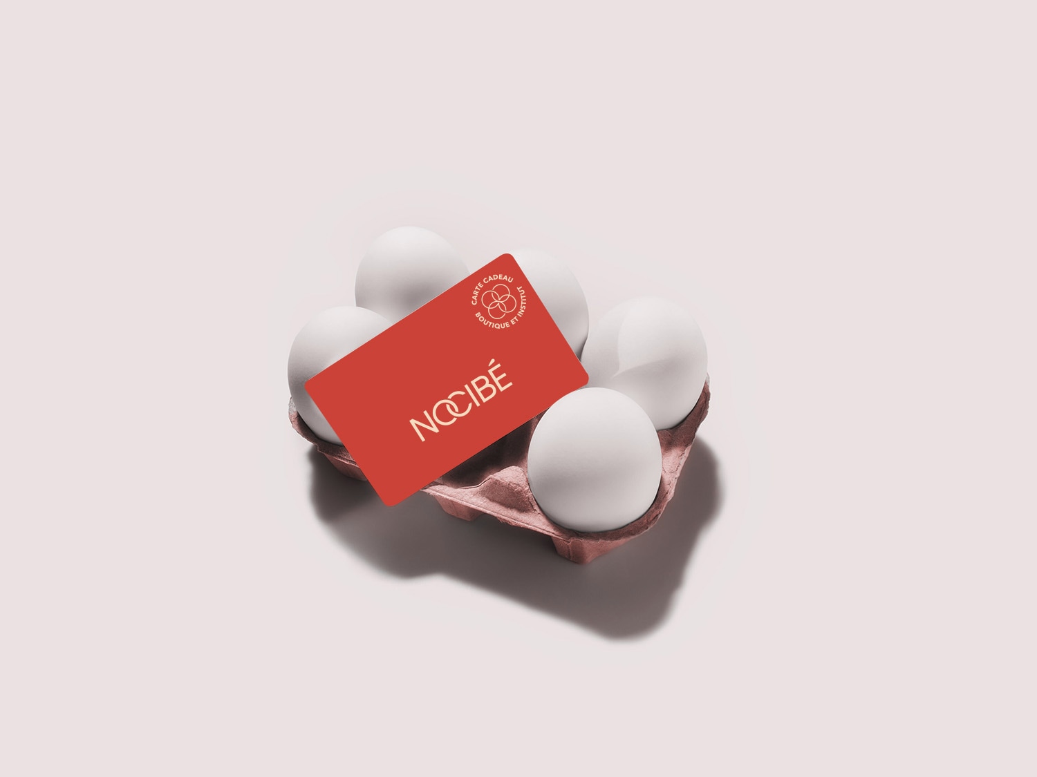 Présentoir à œufs rose pâle avec des œufs blancs et une carte cadeau Nocibé rouge.